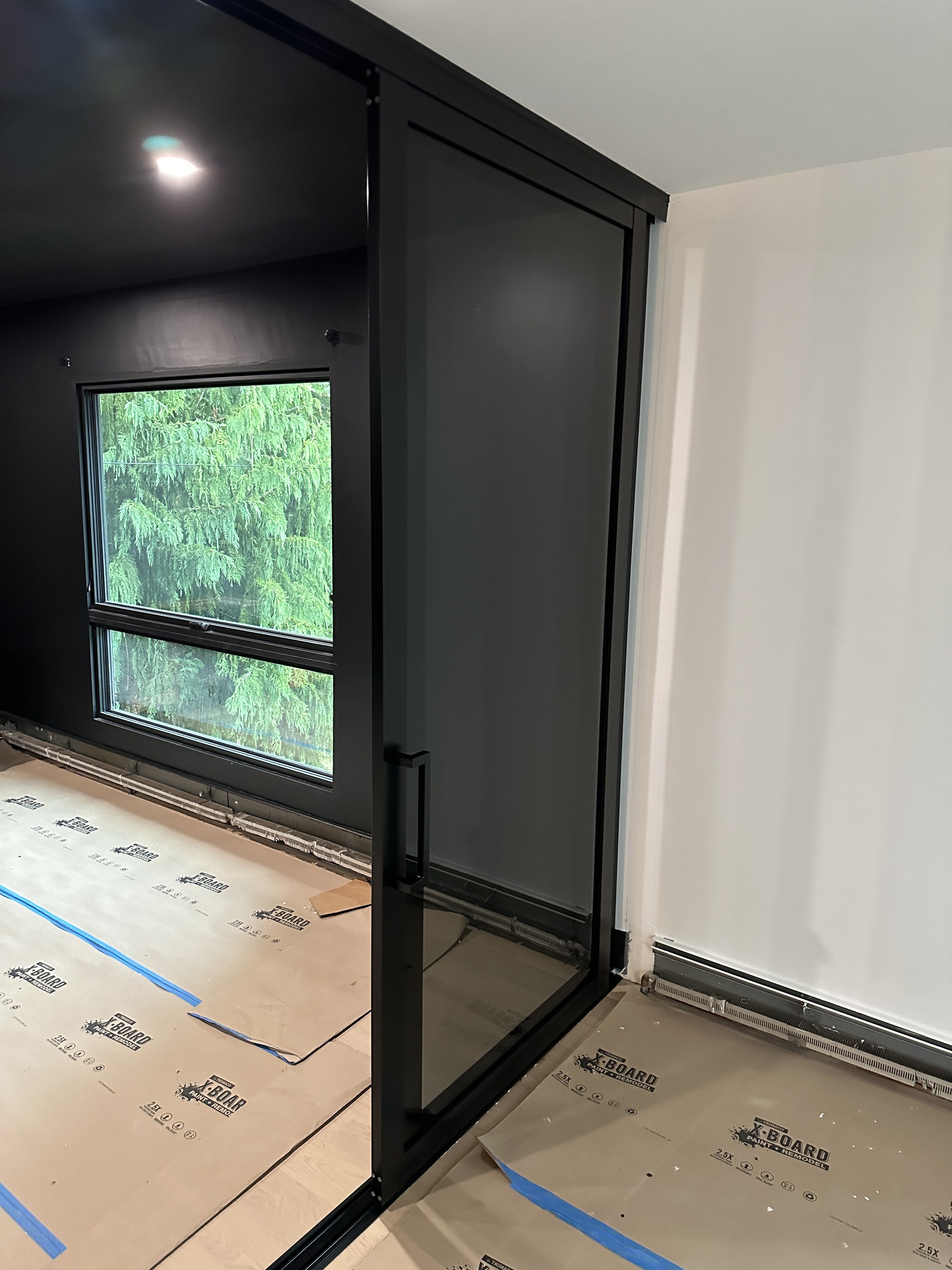 Black steel-framed glass wall and pivoting door during installation, interior view with a window and floor protection still down