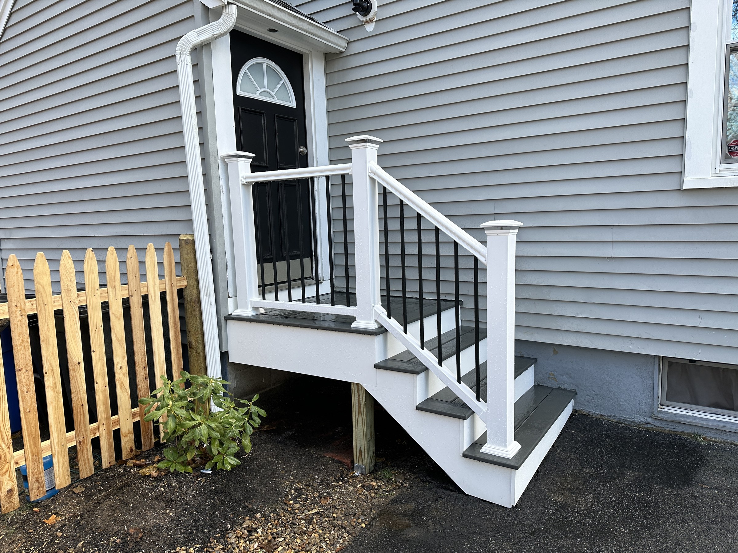 Finished entry steps viewed from the side, white PVC railing with black balusters and dark composite treads, new wood picket fence and a small shrub alongside, Reading
