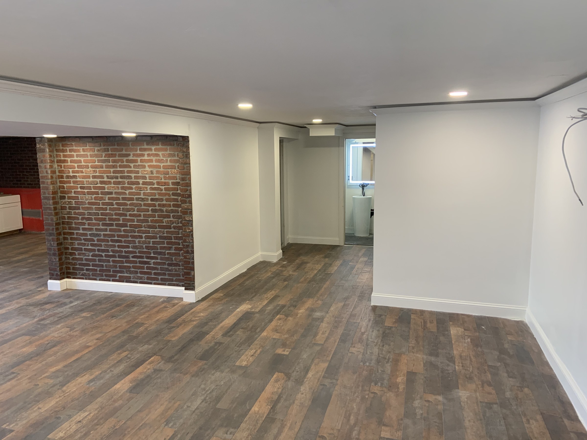 Basement main room with brick accent wall repointed and wide-plank flooring down, sightline through to an ensuite half bath; one ceiling wire at upper right is still unterminated