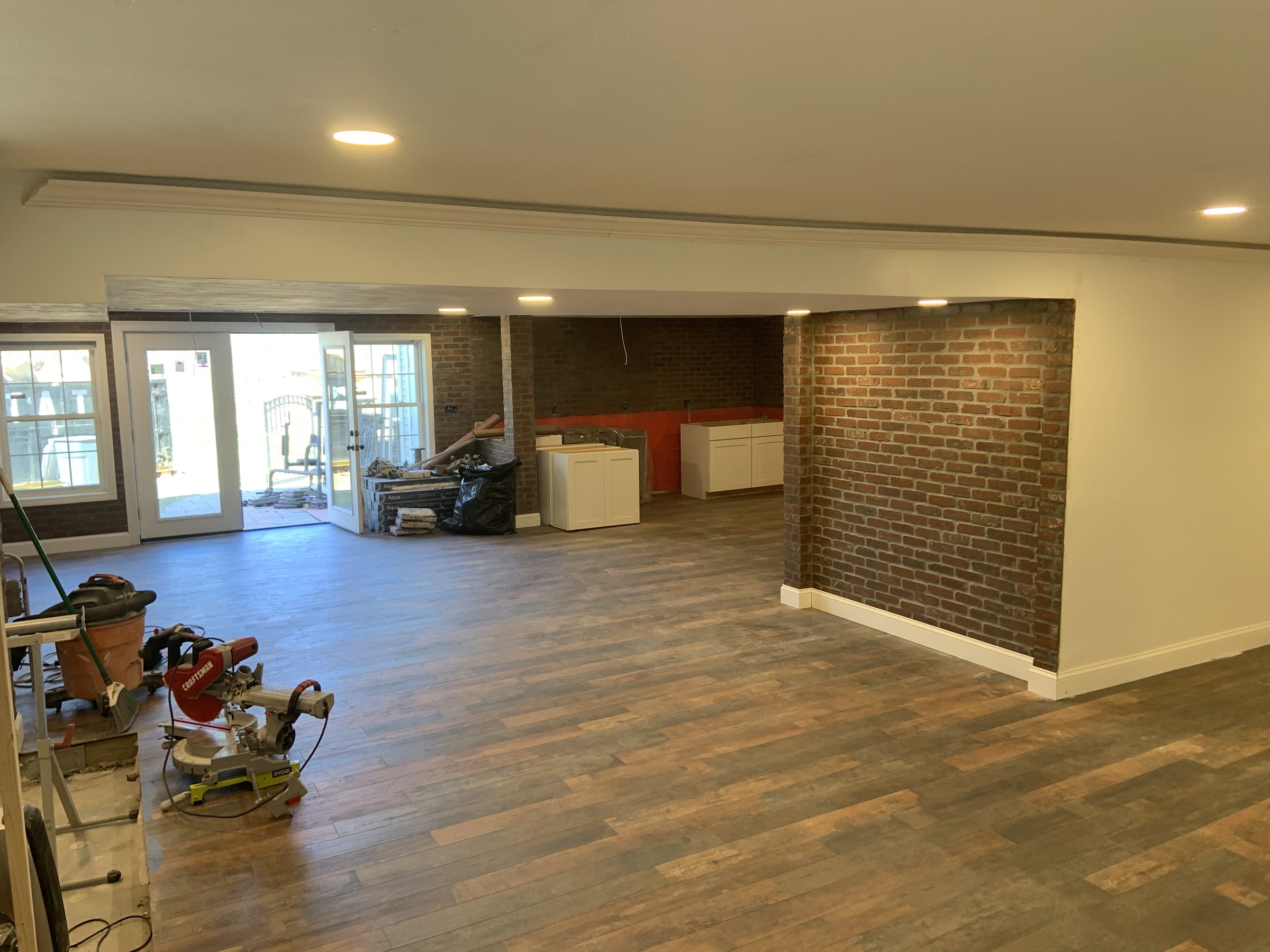 The same basement mid-renovation: brick pillar not yet repointed, flooring and cabinet boxes staged, miter-saw station set up