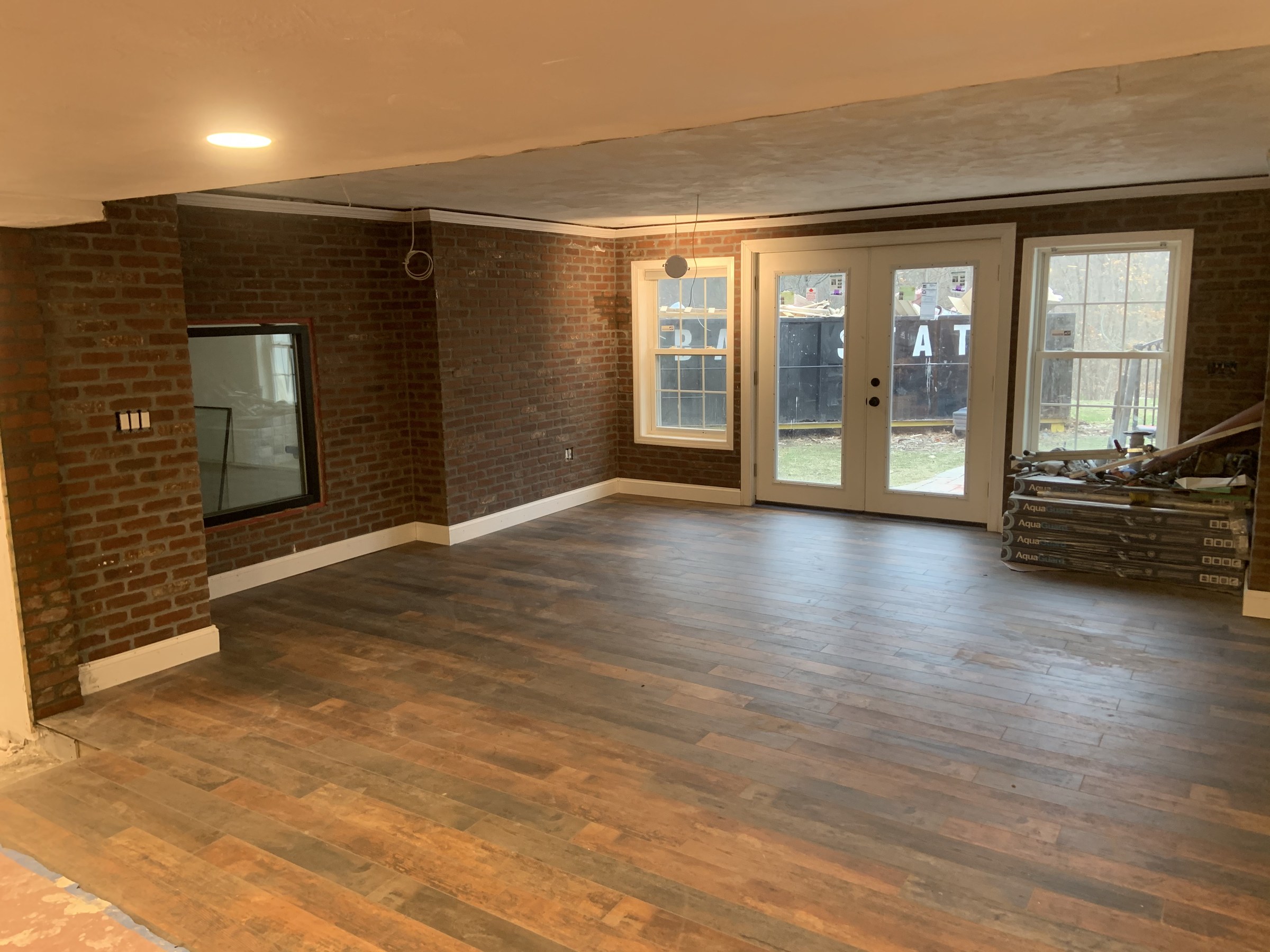 Basement corner during renovation: brick not yet repointed, a second glazed window, and white French doors out to the yard, flooring boxes staged for the next room