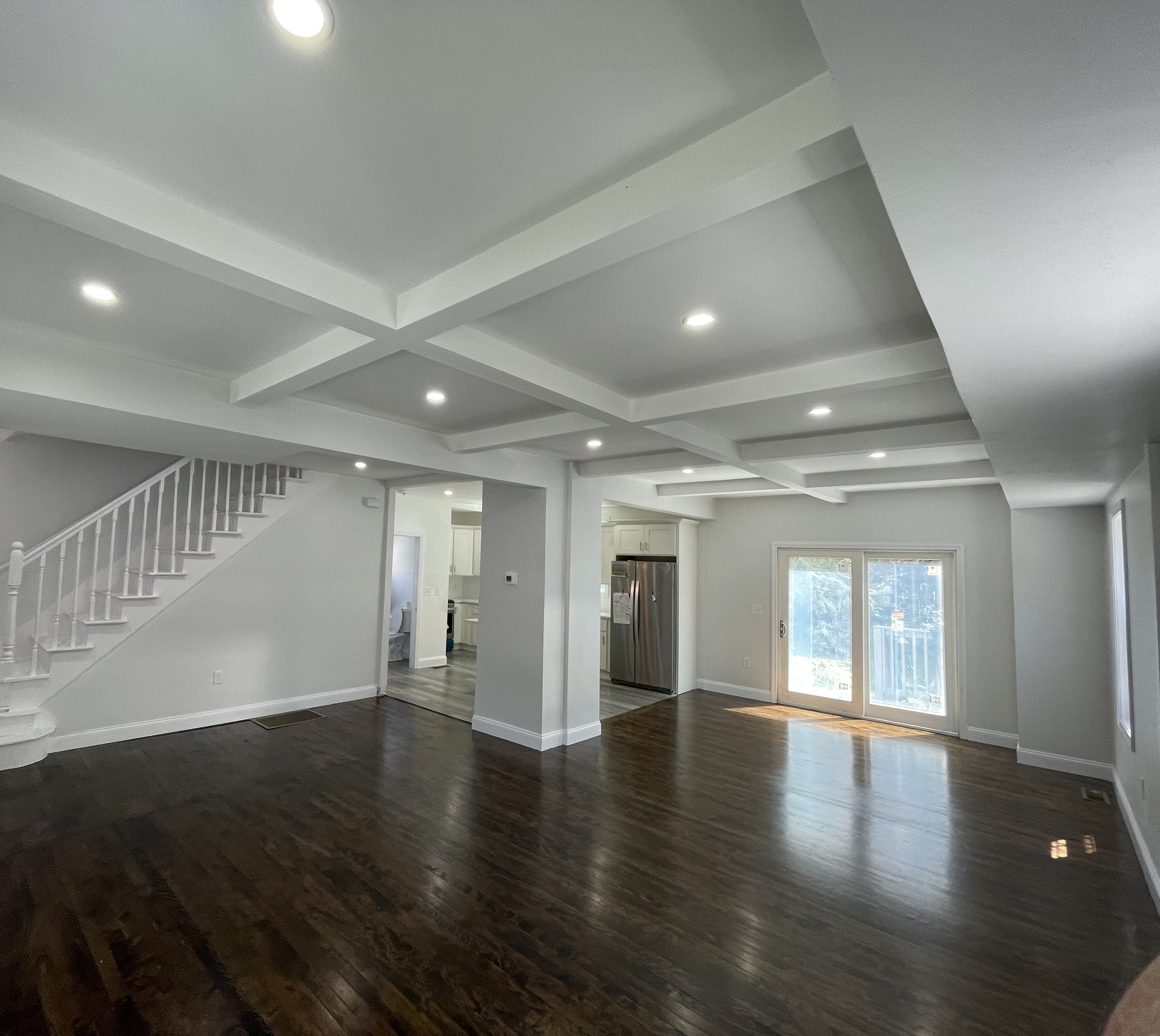 Wide-angle view of a great room with a coffered ceiling and a curved white staircase with turned balusters, a sliding glass door and a glimpse of the kitchen and a powder room beyond
