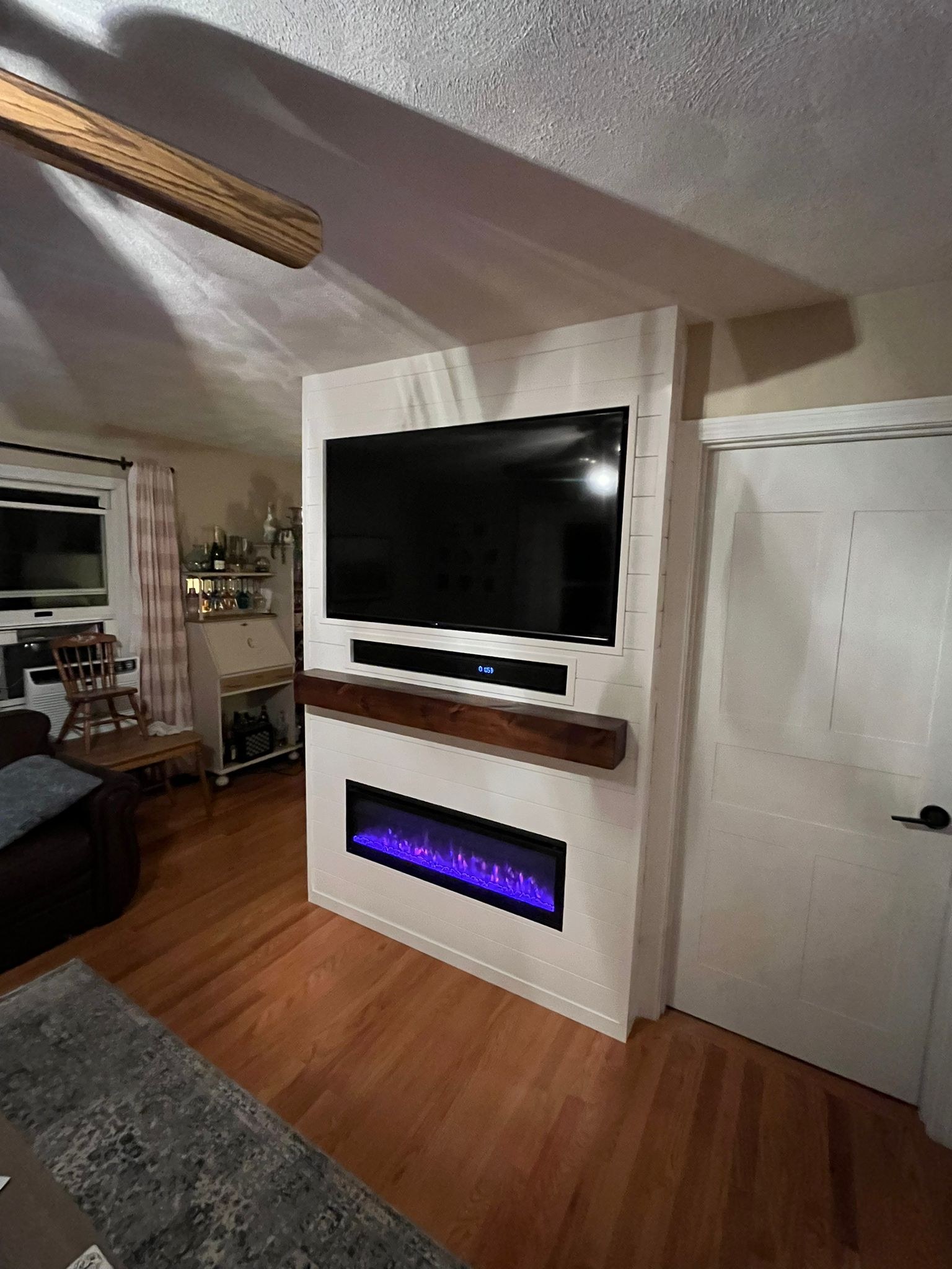 Finished shiplap fireplace wall with a rough-hewn wood mantel, a recessed TV niche above and an electric fireplace insert below, bar cart and room visible alongside